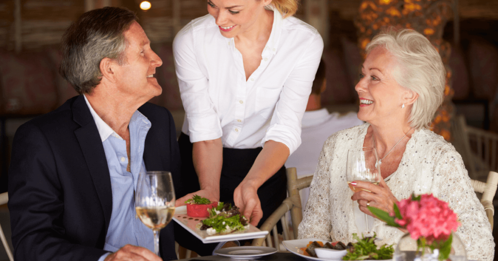 Senior couple out at dinner with waitress serving them food. Enhanced Living