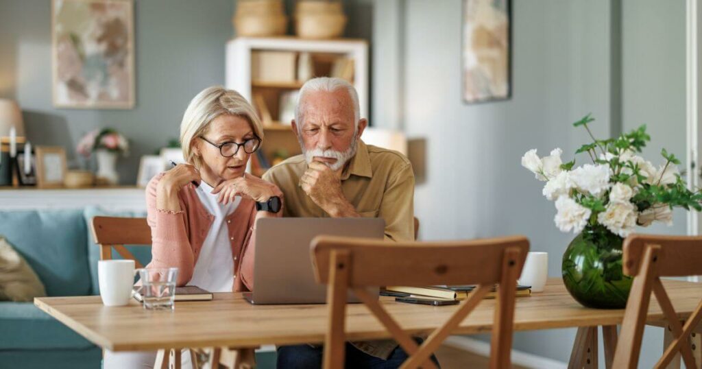 Senior couple sitting at the kitchen table at a laptop comparing the cost of assisted living.